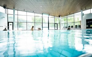 Innenansicht der Siebentäler Therme in Bad Herrenalb. Durch große Panoramafenster fällt viel Tageslicht in die Badehalle mit einer Holzdecke. Im Vordergrund ist das Wasser eines weitläufigen Schwimmbeckens zu sehen, in dem mehrere Badegäste entspannt verweilen. Im Hintergrund stehen Liegestühle vor den Fenstern.