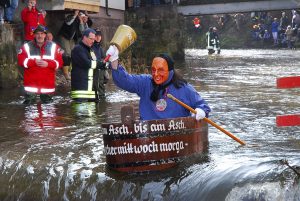 Mit einem blauen Hemd, schwarzem Schal, schwarzer Mütze und Holzmaske im Gesicht, fährt ein Fasnetnarr den Fluss hinab, in der rechten Hand eine hoch erhobene Glocke und in der linken einen Stab, mit dem er steuert. Er ist kurz vor einer kleinen Stromschnelle. Im Hintergrund ein Feuerwehrmann, der bei einer Kenterung helfen würde. Auf dem Zuber steht ein Text, der daran erinnert, dass die Fasnet bis Aschermittwoch geht.