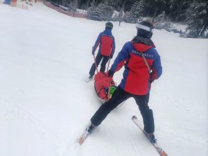Zwei Rettungskräfte der Bergwacht auf Skier transportieren am Notschrei bergabfahrend eine im Rettungsschlitten liegende verletzte Person zum nächsten Versorgungspunkt.