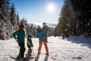 Familie mit zwei Kindern mit Ski- Skimontur macht oben bei sonnigem Wetter am Hang Pause. Ein Kind hat der Vater auf Arm. Hinter ihnen die von Wald begrenzte Abfahrt