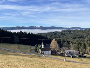 Inmitten der Schwarzwaldlandshaft steht ein kleines Kirchlein mit Nebengebäude. Der Blick streift über die Schwarzwaldberge hinauf zum Feldberg, wo die Berge im Gegnsatz zum tal noch schneebeckt sind.