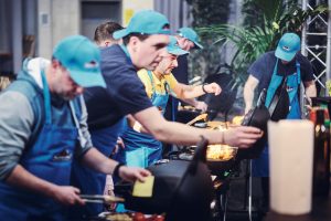 Mehrere Männer stehen gebeugt nebeneinander vor ihrem jeweiligen Grill und grillen um die Wette.