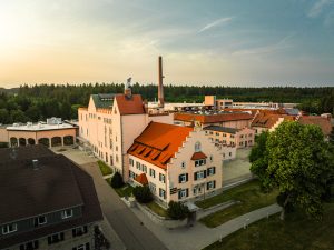 Blick auf die weitläufigen Gebäude einer Brauerei. Im Vordergrund die Brauerei-Gaststätte. die andern Gebäude sind Produktionsstandorte. Überragt werden die Häuser mit ihren gelblichen Wänden und roten Dächern von einem hoch in den Himmel ragenden Kamin.