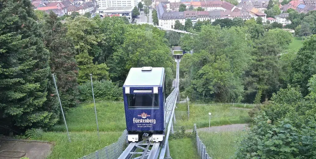 Blick auf die schienengebundene Schlossberg-Bahn mit einer blauen Kabine. Der Blick geht über die Bäume weit nach unten über die Stadt und zum Ende des Gleises an dessen Ende die Talstation sichtbar wird