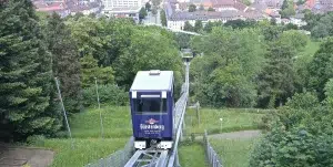 Blick auf die schienengebundene Schlossberg-Bahn mit einer blauen Kabine. Der Blick geht über die Bäume weit nach unten über die Stadt und zum Ende des Gleises an dessen Ende die Talstation sichtbar wird
